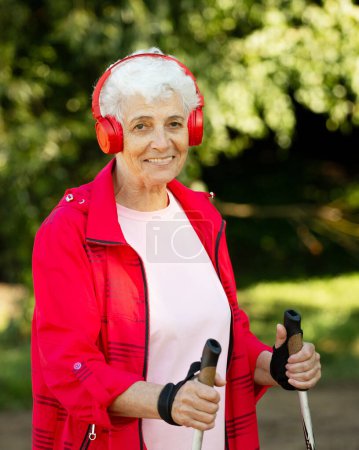 Téléchargez les photos : Une femme souriante âgée entraîne l'endurance lors de la marche nordique. Vieille musique d'écoute féminine dans les écouteurs. - en image libre de droit