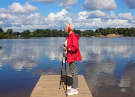 Téléchargez les photos : Une femme âgée aux cheveux courts, vêtue d'une veste rouge, se tient debout avec des bâtons de marche nordique sur un pont au bord du lac et écoute de la musique avec des écouteurs. - en image libre de droit