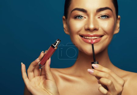 Téléchargez les photos : Une belle jeune femme tient du rouge à lèvres près de son visage sur fond bleu, fait du maquillage, le concept de visage et de cosmétiques. - en image libre de droit