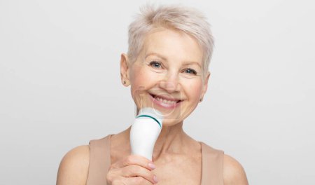 Téléchargez les photos : Femme âgée souriante aux cheveux courts utilisant un masseur facial pour une peau fraîche parfaite - en image libre de droit