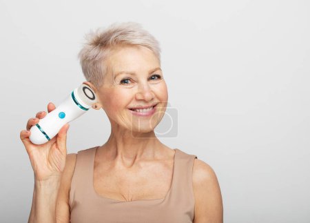 Téléchargez les photos : Femme âgée souriante aux cheveux courts utilisant un masseur facial pour une peau fraîche parfaite - en image libre de droit