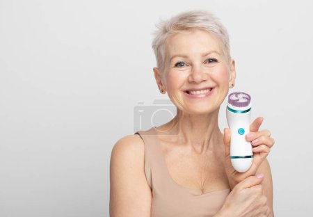 Foto de Mujer anciana sonriente con cabello corto usando masajeador facial para una piel fresca perfecta - Imagen libre de derechos