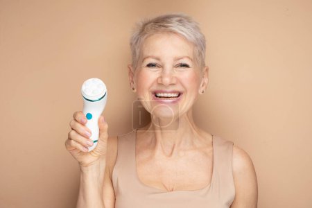 Téléchargez les photos : Femme âgée souriante aux cheveux courts utilisant un masseur facial pour une peau fraîche parfaite sur fond beige - en image libre de droit