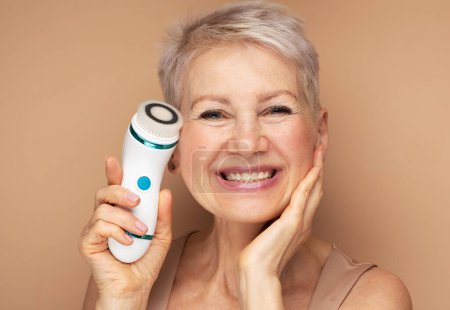 Lächelnde ältere Frau mit kurzen Haaren mit Gesichtsmassagegerät für perfekte frische Haut auf beigem Hintergrund