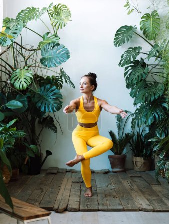 Téléchargez les photos : Brunette femme âgée pratique le yoga à la maison parmi les fleurs. jungle urbaine et gymnastique, yoga, pilates. - en image libre de droit