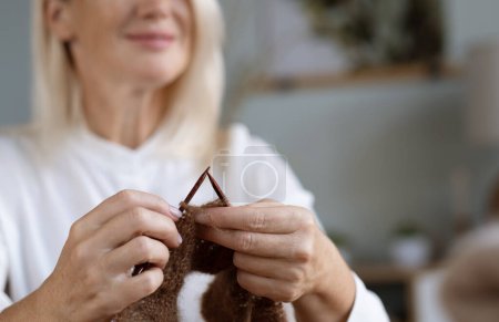 Téléchargez les photos : Pour mes enfants. Heureuse femme âgée enchantée tenant des aiguilles à tricoter et souriant tout en faisant une écharpe - en image libre de droit