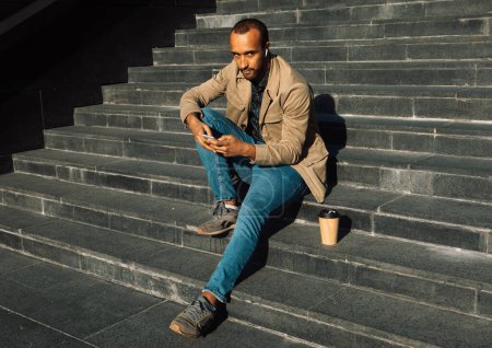 Téléchargez les photos : Portrait de jeune homme afro-américain beau assis sur les marches en utilisant mobile - en image libre de droit