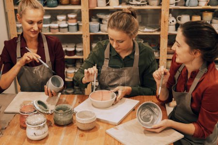 Foto de Una compañía de tres alegres amigas jóvenes están pintando cerámica en un taller de cerámica. Concepto de estilo de vida y personas. - Imagen libre de derechos
