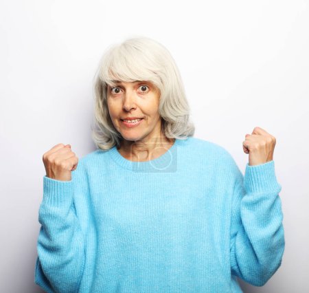 an elderly gray-haired woman in a blue sweater clenches her fists