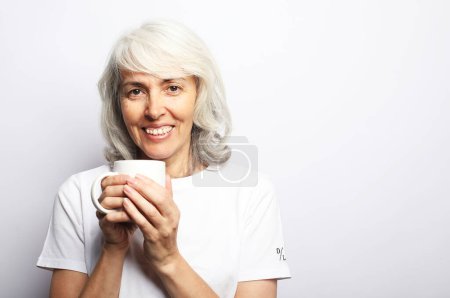 Téléchargez les photos : Une femme âgée aux cheveux gris dans un pull bleu tient une tasse blanche de thé ou de café sur fond gris - en image libre de droit