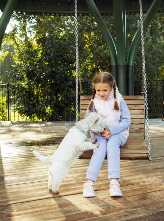Téléchargez les photos : Concept de style de vie et d'amitié. Petite fille blonde monter sur une balançoire avec un chien blanc. - en image libre de droit