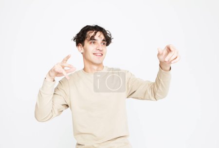Photo for A cheerful smiling young man in a beige sweater dances on a white studio background, enjoys life - Royalty Free Image