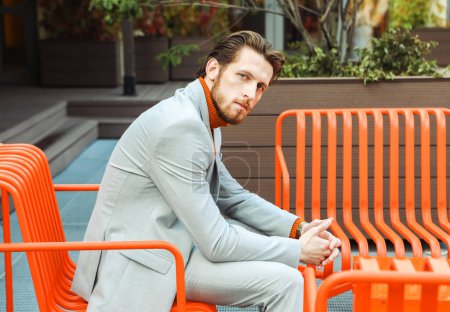 Téléchargez les photos : Jeune homme élégant à col roulé orange et costume à la mode assis sur un banc un jour d'été - en image libre de droit