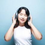 Pretty smiling Asian teenage girl listens to music on headphones over blue background. Close up portrait.