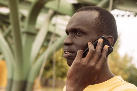 Foto de Un hombre de piel oscura con una camiseta amarilla habla por teléfono un día de verano en el parque. Concepto de estilo de vida y personas. - Imagen libre de derechos
