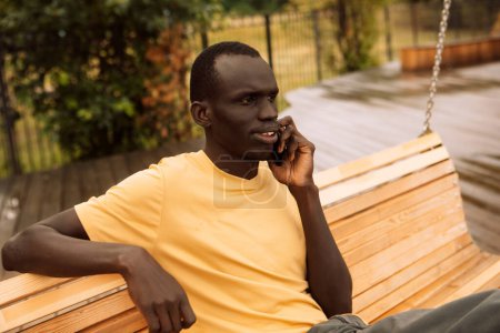 Téléchargez les photos : Un homme à la peau foncée en T-shirt jaune parle au téléphone un jour d'été dans le parc. Style de vie et concept de personnes. - en image libre de droit