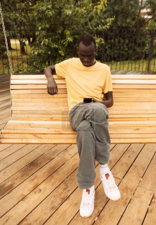 Téléchargez les photos : Jeune homme afro-américain à la peau foncée assis sur une balançoire dans le parc un jour d'été, tenant un smartphone et envoyant des SMS ou parcourant les nouvelles sur les réseaux sociaux. - en image libre de droit