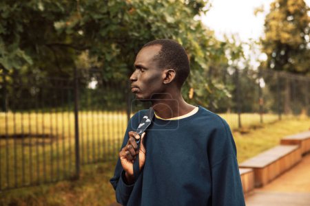 Téléchargez les photos : Portrait d'un jeune homme africain beau vêtu d'un sweat-shirt bleu, tenant un sac à dos, marchant dans le parc un jour d'été - en image libre de droit