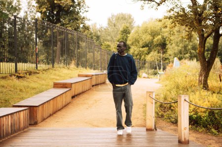 Foto de Retrato de un joven guapo africano vestido con una sudadera azul, sosteniendo una mochila, caminando por el parque en un día de verano - Imagen libre de derechos