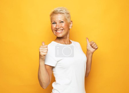 Foto de Senior mujer feliz vistiendo camiseta blanca mostrando pulgares hacia arriba sobre fondo amarillo. - Imagen libre de derechos