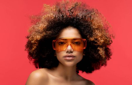 Téléchargez les photos : Jeune femme afro portant des lunettes de soleil orange souriant, isolée sur fond rouge studio. Portrait en gros plan. - en image libre de droit