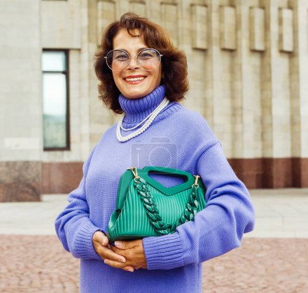 Téléchargez les photos : Une dame âgée joyeuse dans un pull violet tenant le sac à main vert se relaxe dans la rue un jour d'été. Style de vie et concept de personnes. - en image libre de droit