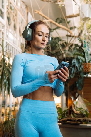 Téléchargez les photos : Femme tenant un smartphone et portant un casque écoute de la musique, se détend pendant Pilates, formation de yoga dans une salle avec des plantes tropicales. - en image libre de droit