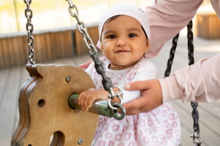 little smiling baby girl sits on a swing in the park on a summer day, time for games