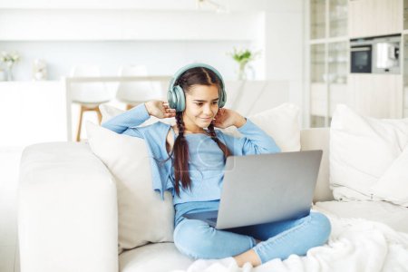 Schoolgirl with pigtails wearing headphones sits on the sofa and works with a laptop