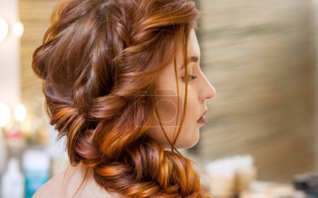 Téléchargez les photos : Belle, aux longs cheveux roux, la coiffeuse tisse une tresse française, dans un salon de beauté. Soins capillaires professionnels et création de coiffures
. - en image libre de droit