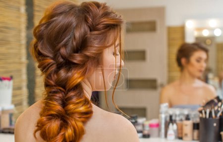 schön, mit langen, rothaarigen haarigen Mädchen, Friseur webt einen französischen Zopf, in einem Schönheitssalon. professionelle Haarpflege und Frisurengestaltung.