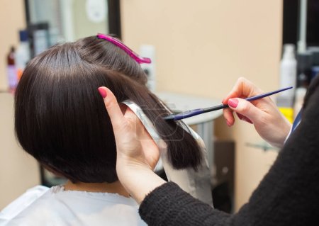 Foto de The hairdresser paints the woman's hair in a dark color, apply the paint to her hair in the beauty salon. - Imagen libre de derechos