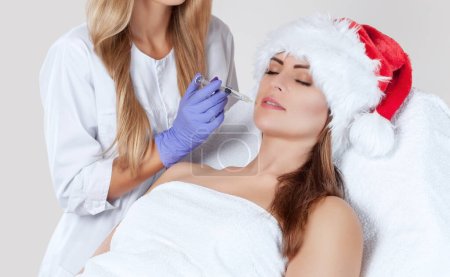 Téléchargez les photos : The doctor cosmetologist makes  injection on the face skin and lips of a beautiful, young woman in the Santa Claus hat. New Year's and Cosmetology concept. - en image libre de droit