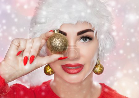 Foto de Retrato de una hermosa mujer con un sombrero rojo de Santa Claus y suéter rojo de punto sosteniendo una bola de navidad dorada sobre fondo de copos de nieve. El concepto de Año Nuevo. - Imagen libre de derechos