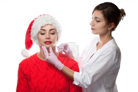 Téléchargez les photos : Le médecin cosmétologue fait une injection sur les lèvres d'une belle femme dans le chapeau du Père Noël. Concept Nouvel An et cosmétologie - en image libre de droit