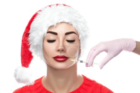Téléchargez les photos : Le médecin cosmétologue fait l'injection de toxine botulique sur la peau du visage d'une belle jeune femme au chapeau de Père Noël. Concept Nouvel An et cosmétologie - en image libre de droit