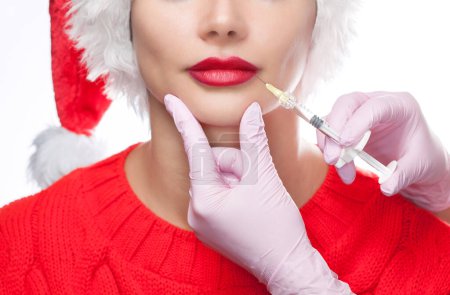 Téléchargez les photos : Le médecin cosmétologue fait une injection sur les lèvres d'une belle femme dans le chapeau du Père Noël. Concept Nouvel An et cosmétologie - en image libre de droit