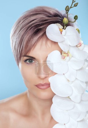Téléchargez les photos : Portrait d'une belle femme dans un salon de spa avec orchidée blanche à la main sur un fond bleu. Soins professionnels de la peau. - en image libre de droit