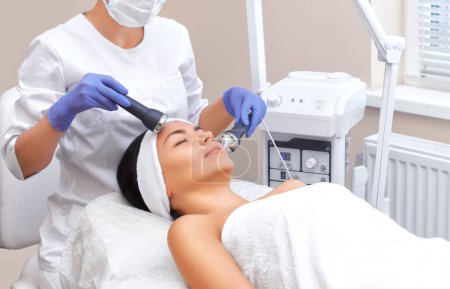 Téléchargez les photos : Le médecin-cosmétologue fait de la procédure un nettoyage ultrasonique de la peau du visage d'une belle jeune femme dans un salon de beauté.. - en image libre de droit