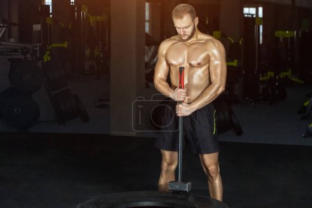 Photo for Sport muscular man hitting wheel tire with hammer sledge in the gym. Sports training - Royalty Free Image