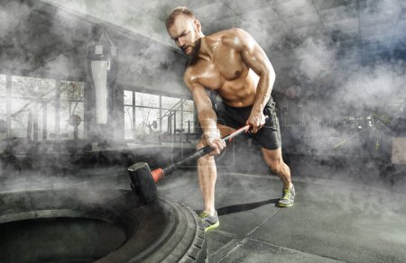 Photo for Sport muscular man hitting wheel tire with hammer sledge in the gym. Sports training - Royalty Free Image