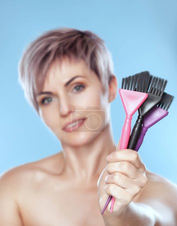 Photo for Portrait of a beautiful blonde woman with beautiful make-up and short haircut after dyeing hair holds in hand brush for dyeing hair in a hairdressing salon on a blue background. - Royalty Free Image