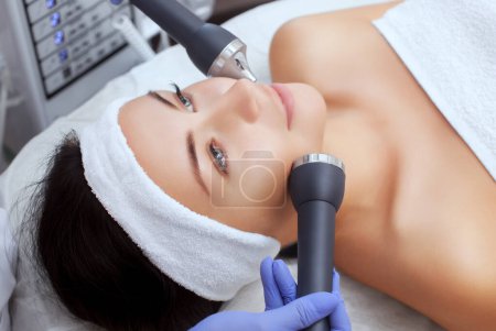 Foto de El cosmetólogo hace del procedimiento una limpieza ultrasónica de la piel facial de una hermosa mujer joven en un salón de belleza. Cosmetología y cuidado profesional de la piel
. - Imagen libre de derechos