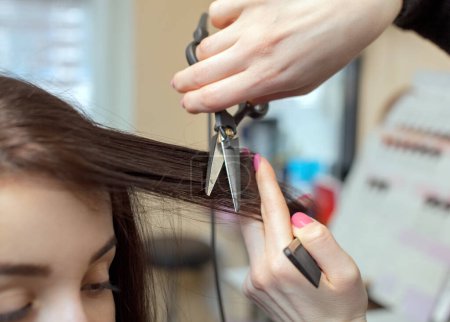 Téléchargez les photos : Le coiffeur fait une coupe de cheveux avec des ciseaux chauds de cheveux à une jeune fille, une brune dans un salon de beauté.. - en image libre de droit