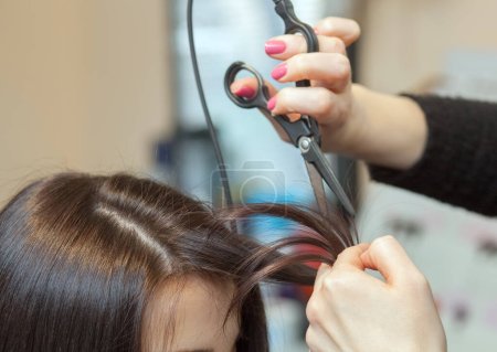 Téléchargez les photos : Le coiffeur fait une coupe de cheveux avec des ciseaux chauds de cheveux à une jeune fille, une brune dans un salon de beauté.. - en image libre de droit