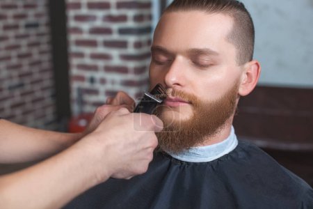 Photo for A male barber at work, he does a beard and mustache haircut to a man in the barbershop. Male cosmetology, care of the beard, creating a stylish look. - Royalty Free Image
