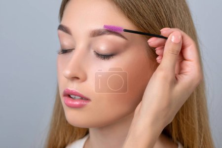 Make-up-Artist kämmt Augenbrauen mit einem Pinsel zu einem schönen jungen blonden Teenie-Mädchen mit langen Haaren, Make-up, saubere Haut nach permanentem Makeup.Make-up-Konzept, Augenbrauenform modellieren.