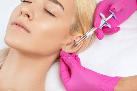 Photo for The doctor cosmetologist makes the injections procedure for smoothing wrinkles and against flabbiness of the skin on earlobe of a beautiful, young woman.Women's cosmetology in the beauty salon. - Royalty Free Image