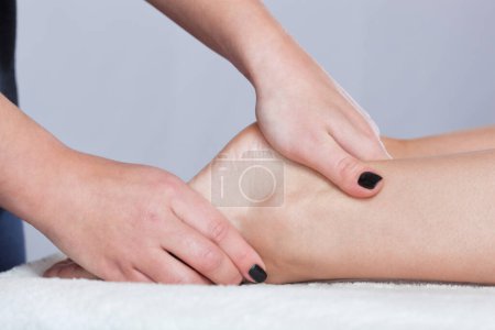 Foto de Un médico ortopédico masajea el pie y la parte inferior de la pierna del paciente después de una lesión Esguince de tobillo en la clínica.Concepto de cosmetología y masaje. - Imagen libre de derechos