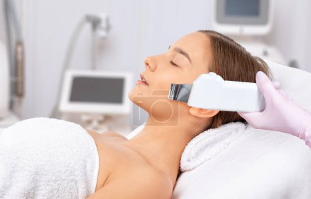 The cosmetologist makes the ultrasound cleaning procedure of the facial skin of a beautiful, young woman in a beauty salon. Cosmetology and professional skin care.